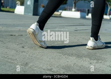 Primo piano dei piedi della donna che camminano su una superficie in cemento, indossando sneakers bianche e leggings neri, sottolineando l'attività urbana e le calzature Foto Stock
