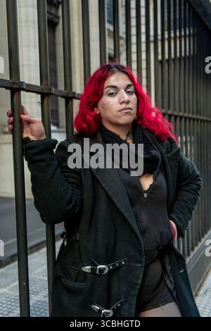 Ritratto di una giovane donna urbana con i capelli rosa e i tatuaggi, appoggiata su una recinzione metallica in città Foto Stock