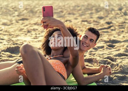 Una bella giovane coppia giace sulla sabbia e sorride felicemente mentre si fa un selfie insieme a un telefono cellulare durante le vacanze estive al mare. Foto Stock