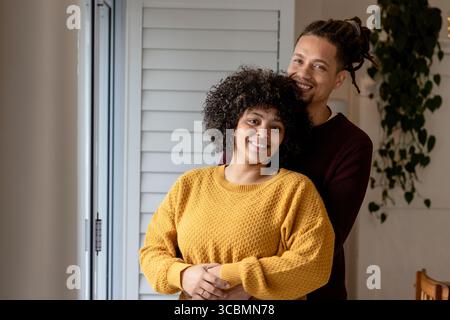 Coppia afroamericana che si abbraccia mentre si trova davanti a una finestra con persiane nel soggiorno con pianta in vaso. Romantico, calore, appagamento, accogliente, sincero, fami Foto Stock