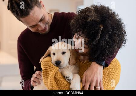 Coppia afroamericana con cucciolo Golden retriever in cucina e maglioni con smartwatch. Famiglia, compagnia, accogliente, domestico, affettuoso Foto Stock