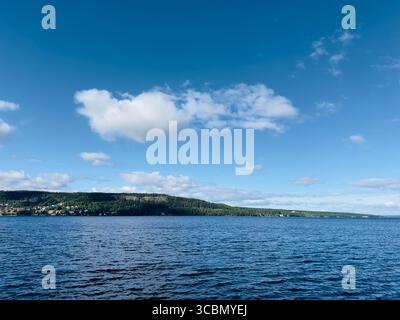 La tranquilla scena del lago mostra un cielo azzurro luminoso con soffici nuvole bianche sopra un lontano Treeline. Foto Stock