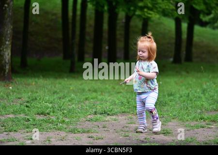 Il bambino dai capelli rossi cammina lungo un sentiero erboso vicino al parco giochi nel soleggiato parco, tenendo in mano un piccolo bastoncino. Foto Stock