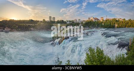 Potenti cascate d'acqua scendono lungo le rocce delle Cascate del Reno, circondate dal verde e dagli edifici cittadini durante il tramonto. Il cielo si illumina con un caldo bagliore come Foto Stock
