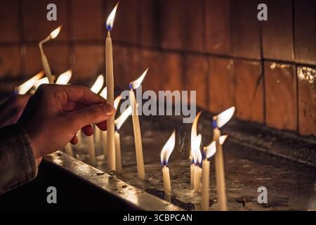 Un primo piano di una mano che posiziona con cura una candela accesa tremolante su un piedistallo, circondato da altre candele accese in un ambiente spirituale scarsamente illuminato Foto Stock