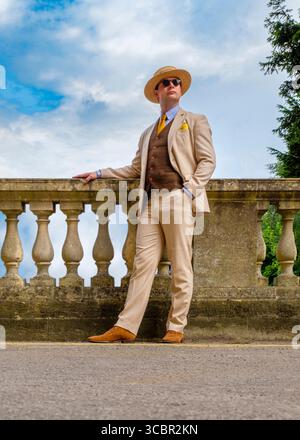 Un uomo con un costume da abbronzatura, un cappello di paglia e occhiali da sole si erge su una sporgenza di pietra, appoggiato su una ringhiera di pietra, con un cielo blu e nuvole sullo sfondo. Foto Stock
