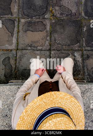 Una persona che indossa un cappello di paglia, un gilet marrone e una tuta beige si siede su una superficie di pietra. Foto Stock