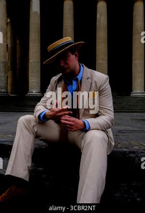 Un uomo con una tuta beige e un cappello di paglia si siede su un gradino di pietra, guardando in basso. Indossa una camicia blu e una cravatta marrone. Lo sfondo presenta un grande colu Foto Stock