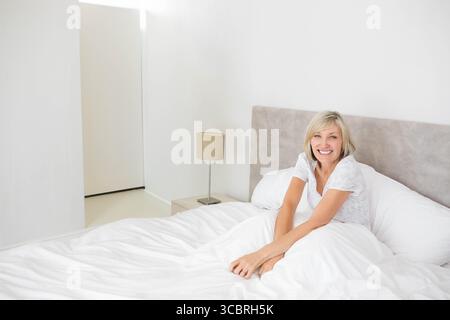 Donna sorridente mentre si siede in posizione eretta sul letto in camera da letto con pigiama con lampada da comodino e spazio fotocopie Foto Stock