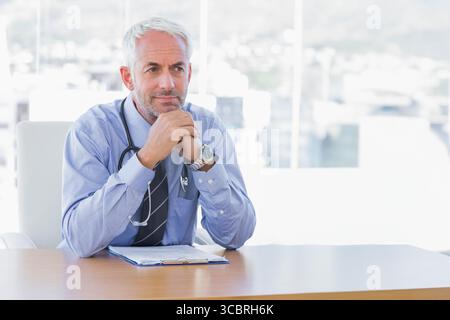 Medico senior che indossa camicia e cravatta appoggiata alla scrivania in ufficio con appunti stetoscopio Foto Stock