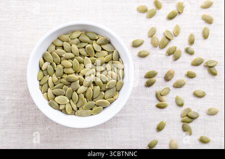 Pepitas, semi di zucca mondati, in una ciotola bianca, su lino. Semi di zucca secchi crudi, frutti di Cucurbita, verde chiaro dopo la rimozione della buccia. Foto Stock