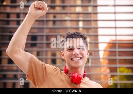 Giovane felice che solleva pugno per festeggiare indossando le cuffie rosse all'aperto Foto Stock