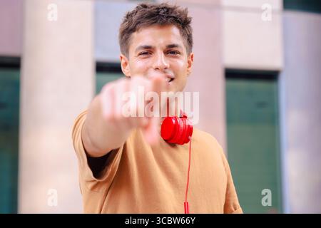 Giovane sorridente che punta alla fotocamera indossando le cuffie rosse all'aperto Foto Stock