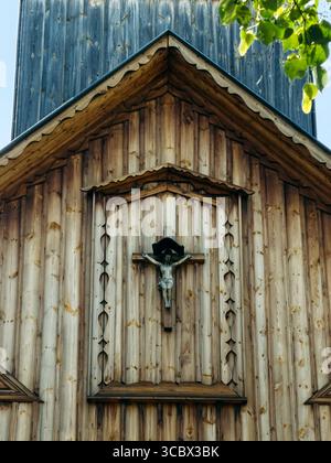 Facciata della chiesa in legno con crocifisso montato sotto un piccolo baldacchino vicino alla cima del tetto. Architettura religiosa, cristianesimo rurale, artigianato in legno Foto Stock