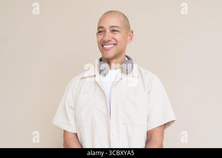 Ritratto di un uomo ispanico calvo che indossa una camicia beige casual e cuffie intorno al collo, appoggiato su un semplice sfondo beige. Foto Stock
