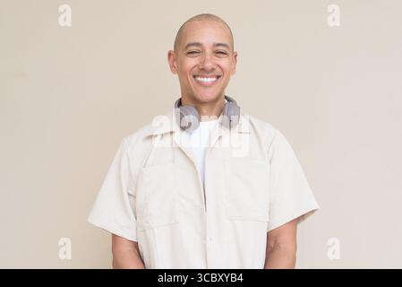 Ritratto di un uomo ispanico calvo che indossa una camicia beige casual e cuffie intorno al collo, appoggiato su un semplice sfondo beige. Foto Stock
