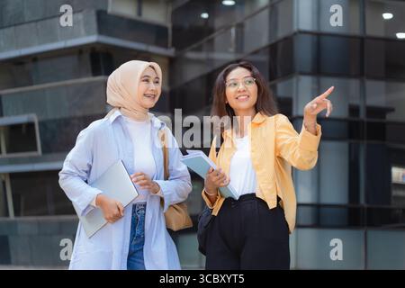 Due donne camminano per strada, una di loro indica qualcosa. Entrambi portano zaini, laptop e una pila di libri. Foto Stock