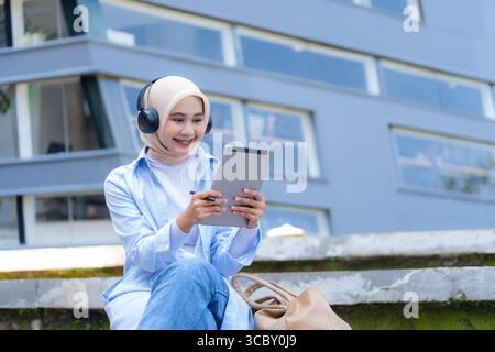 Una donna che indossa una sciarpa per la testa è seduta su una sporgenza con un tablet in mano. Sta sorridendo e si sta godendo il suo tempo Foto Stock