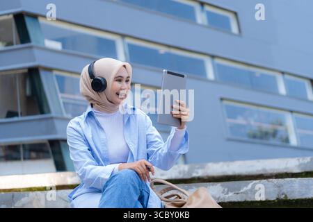 Una donna che indossa una sciarpa per la testa è seduta su una sporgenza con un tablet in mano. Sta sorridendo e si sta godendo il suo tempo Foto Stock
