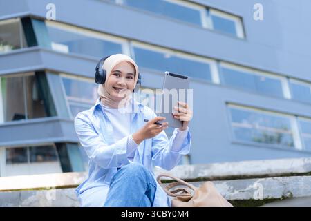 Una donna che indossa una sciarpa per la testa è seduta su una sporgenza con un tablet in mano. Sta sorridendo e si sta godendo il suo tempo Foto Stock