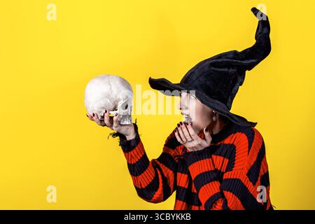 Tema Halloween. Giovane strega in maglione a righe, cappello nero con teschio su sfondo giallo Foto Stock