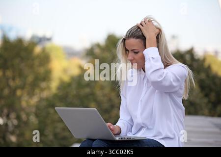 GIOVANE DONNA BIONDA CHE FA NETWORKING SUL SUO NOTEBOOK ALL'APERTO Foto Stock