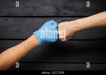 Una mano in un guanto medico blu e una mano femminile senza guanto bump pugni su uno sfondo di legno nero Foto Stock