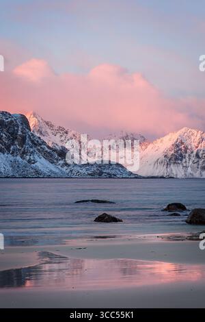 Colorata alba invernale a Haukland Beach, Isole Lofoten, Norvegia. Le montagne innevate e le nuvole rosa si riflettono nel mare calmo e nella sabbia bagnata Foto Stock