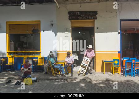 Shiner di scarpe e clienti maschili seduti su mobili colorati di una terrazza e socializzando di fronte a una caffetteria di fronte a una caffetteria Foto Stock