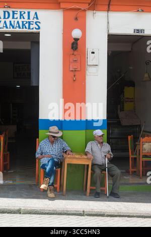 Due uomini anziani seduti ad un tavolino all'ombra di fronte ad una parete colorata di un bar/ristorante Foto Stock