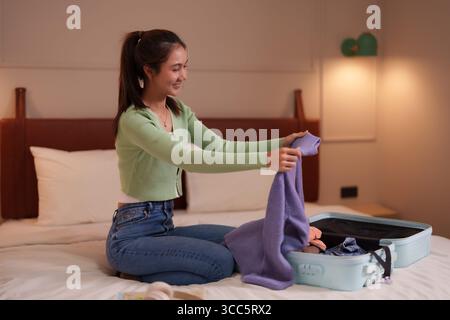 Giovane donna asiatica sorridente vestiti pieghevoli che organizza i suoi bagagli per le vacanze o i viaggi d'affari in una moderna camera da letto pianifica l'avventura in anticipo Foto Stock
