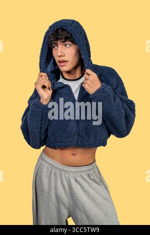 Giovane uomo alla moda che indossa una giacca con cappuccio blu e pantaloni felpa grigi, in posa su sfondo giallo Foto Stock