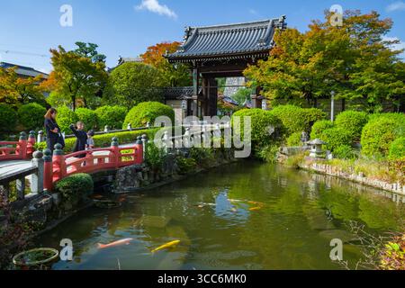 Un tranquillo giardino giapponese caratterizzato da un tranquillo laghetto di koi, vegetazione lussureggiante e elementi architettonici tradizionali. Visitatori che passeggiano su un ponte rosso, e. Foto Stock