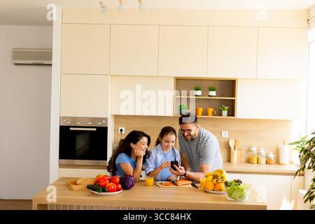 Tre amici si riuniscono in una cucina luminosa e contemporanea, condividendo risate e sorrisi mentre guardano uno smartphone. Il piano di lavoro è decorato con colo Foto Stock