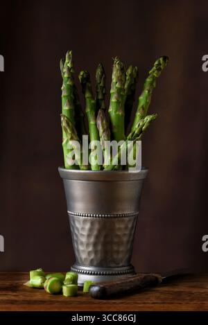 Una tazza di peltro riempita di lance di asparagi su un tavolo in legno rustico e un caldo sfondo a chiazze. Foto Stock