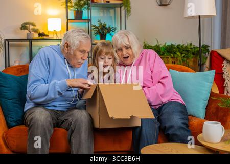 I nonni caucasici felici e contenti e la nipotina che aprono insieme la scatola di cartone si siedono sul divano di casa. I clienti della famiglia sorridenti disimballano un buon pacco guardando all'interno un ottimo acquisto consegnato. Foto Stock