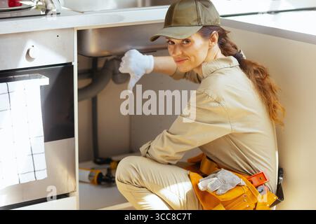 Un idraulico dà il pollice sotto un lavandino della cucina, indicando un problema idraulico. L'immagine suggerisce problemi di riparazione a domicilio e una valutazione negativa. Foto Stock