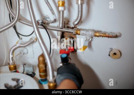 Un primo piano cattura una mano con i guanti che gira una valvola rossa in mezzo a una rete di tubi metallici flessibili, raccordi in ottone e cablaggi, illustrando la casa concentrata Foto Stock