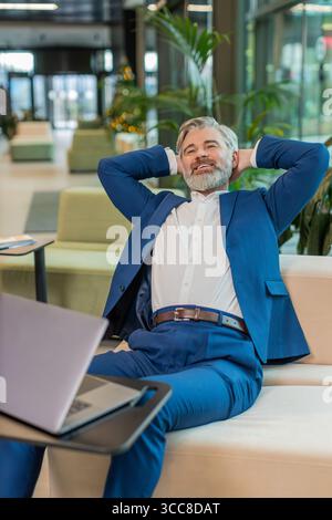 Uomo d'affari felice seduto comodamente con la mano dietro la testa nella moderna hall dell'ufficio chiudendo il computer portatile dopo aver finito il lavoro. Uomo barbuto in tuta che lavora online, e-learning, naviga su Internet su netbook Foto Stock