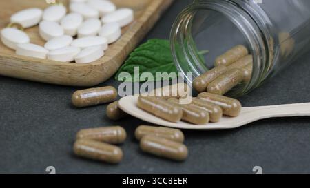 Capsule integratore a base di erbe versate da vaso di vetro con compresse bianche su cucchiaio, concetto di salute naturale e benessere. Foto Stock