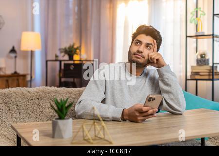 Giovane indiano seduto sul divano a tavola, scrivendo attivamente sullo smartphone, appoggiando la testa a mano nel soggiorno di casa. L'ispanico sembra concentrato, profondamente impegnato in conversazione o attività. Foto Stock