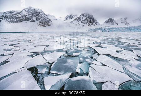 Frantumazione del ghiaccio marino del Circolo Polare Artico e impacchi di ghiaccio sulla costa nord-occidentale delle Svalbard con aspre montagne sullo sfondo Foto Stock