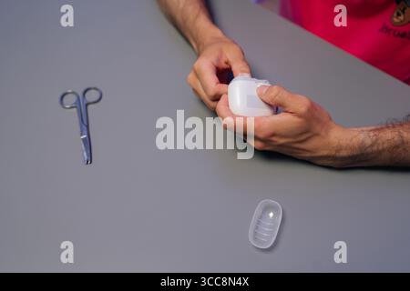 Vista dall'alto dell'uomo che utilizza il regolacapelli elettrico per la manicure domestica, circondato da forbici per unghie e contenitore su un tavolo grigio, concentrandosi sul personale Foto Stock