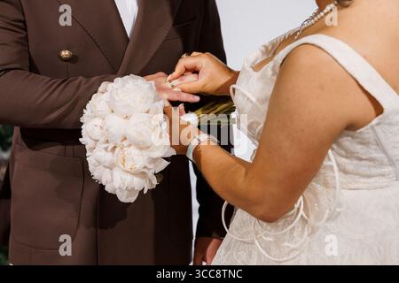 Un primo piano cattura uno scambio di anelli nuziali, mostrando uno sposo con una tuta marrone posizionando una fascia sul dito della sposa, il bouquet visibile Foto Stock