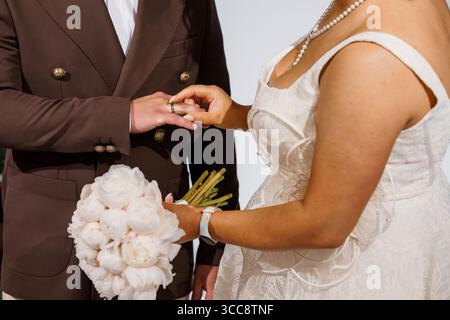 Uno scatto ravvicinato che cattura il momento toccante di uno scambio di anelli nuziali tra uno sposo con un abito marrone e una sposa con un abito in pizzo bianco Foto Stock