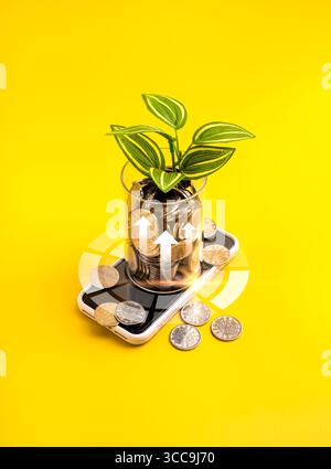 Vaso di vetro con monete e piante che crescono su smartphone su sfondo giallo verticale, concetto di crescita finanziaria, investimento digitale, tecnologia fintech, mobile Foto Stock