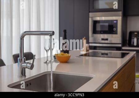 Cucina moderna a isola con rubinetto cromato con acqua corrente, piano cottura a induzione e accessori eleganti in uno spazio ben illuminato. Foto Stock