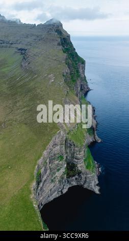 Le scogliere torreggianti delle Isole Faroe si estendono drammaticamente su acque calme e profonde. Lussureggianti colline verdi si fondono con aspre formazioni rocciose, creando una vista mozzafiato e tranquilla sulla costa. Foto Stock