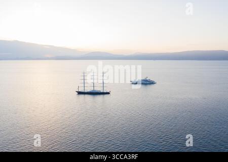 Idra, Grecia - 30 luglio 2025: Veduta aerea di due maestosi yacht che attraversano le tranquille e profonde acque blu sotto il soffice bagliore di un'alba serena. Foto Stock