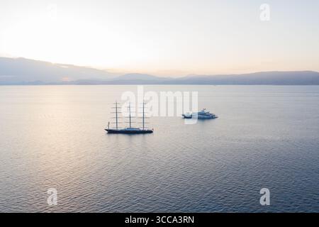 Idra, Grecia - 30 luglio 2025: Vista aerea del mare sereno che riflette il soffice bagliore dell'alba, culla una maestosa barca a vela e un elegante yacht in un abbraccio tranquillo. Foto Stock